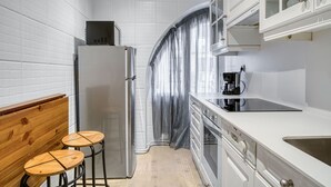 Une cuisine compacte avec des meubles blancs, un réfrigérateur en acier inoxydable, une machine à café et deux tabourets en bois.