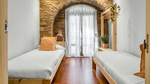 Une chambre avec deux lits simples, un mur en pierre, une grande fenêtre avec des rideaux et un miroir sur le mur.