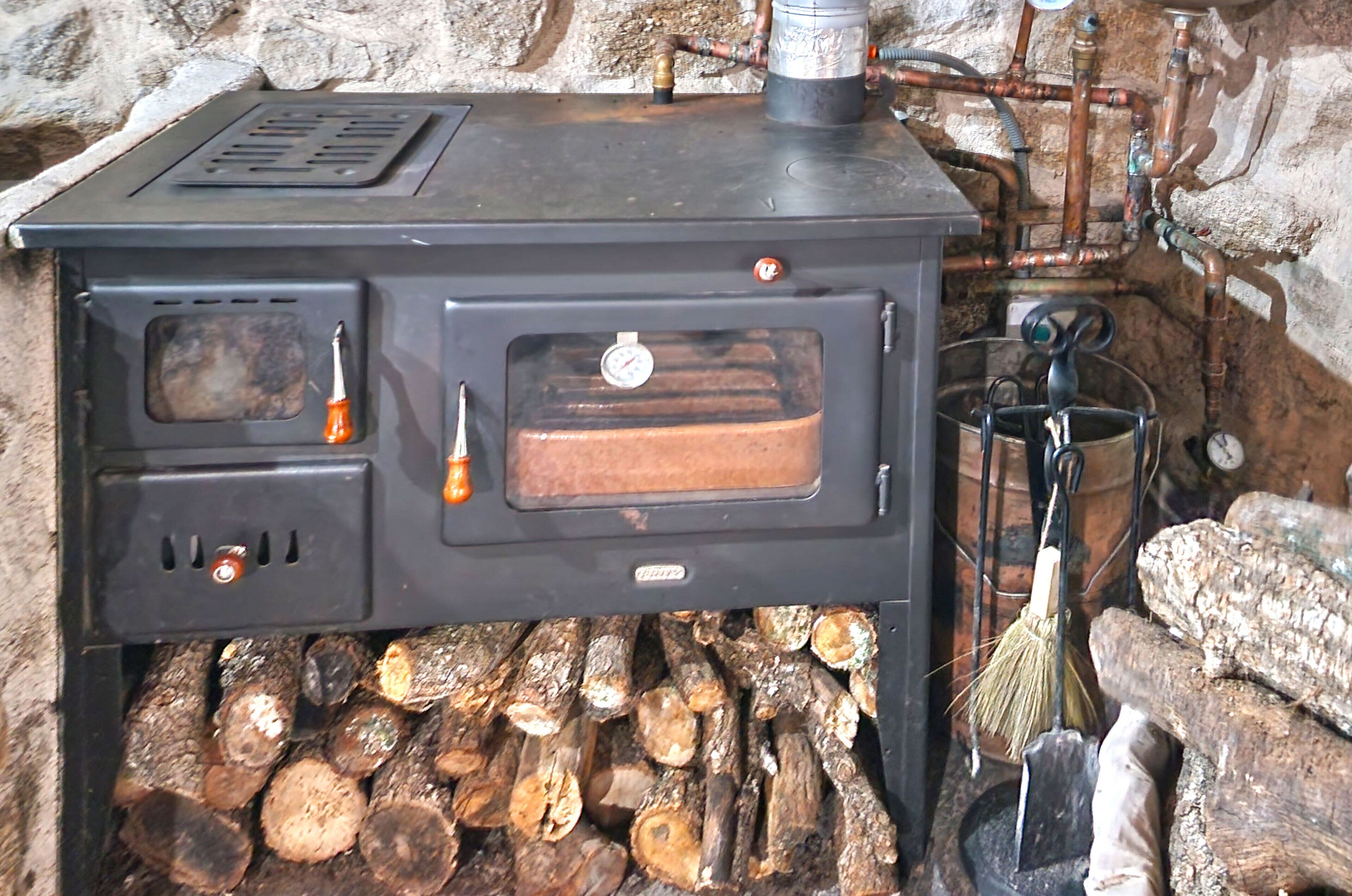 Un poêle à bois muni d’un thermomètre, avec un récipient en métal placé sur le dessus et du bois de chauffage rangé en dessous.