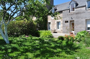 A stone house with a garden in front, a tree, and a clear blue sky.