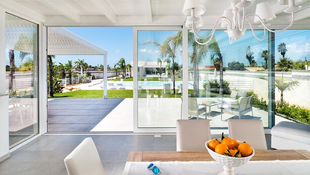 A modern room with a glass wall, a dining table with a bowl of fruit, and a view of a pool and greenery outside. - Villa with private pool, 300 metres from beach - DiCasaInSicilia (Marsala)
