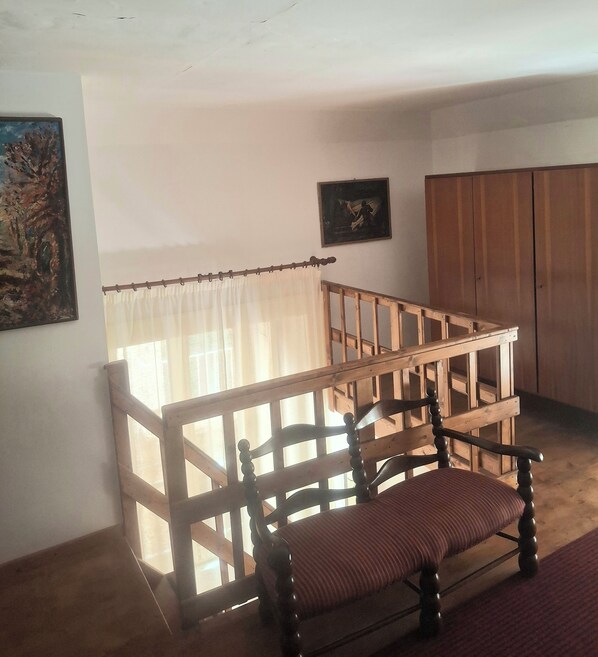 Een kamer met een houten stoel, een houten bank, een raam met gordijnen, een schilderij aan de muur en een kledingkast.