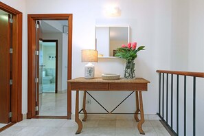Ein Flur mit Holztür, einem Spiegel, einem Tisch mit Lampe, einer Vase mit Blumen und einem Geländer.