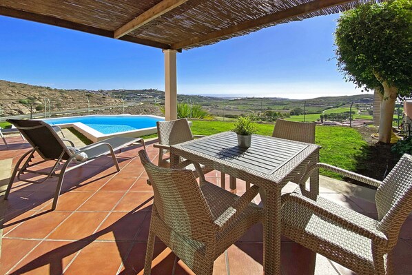 A covered outdoor area with a pool, wicker furniture, and a view of the countryside.