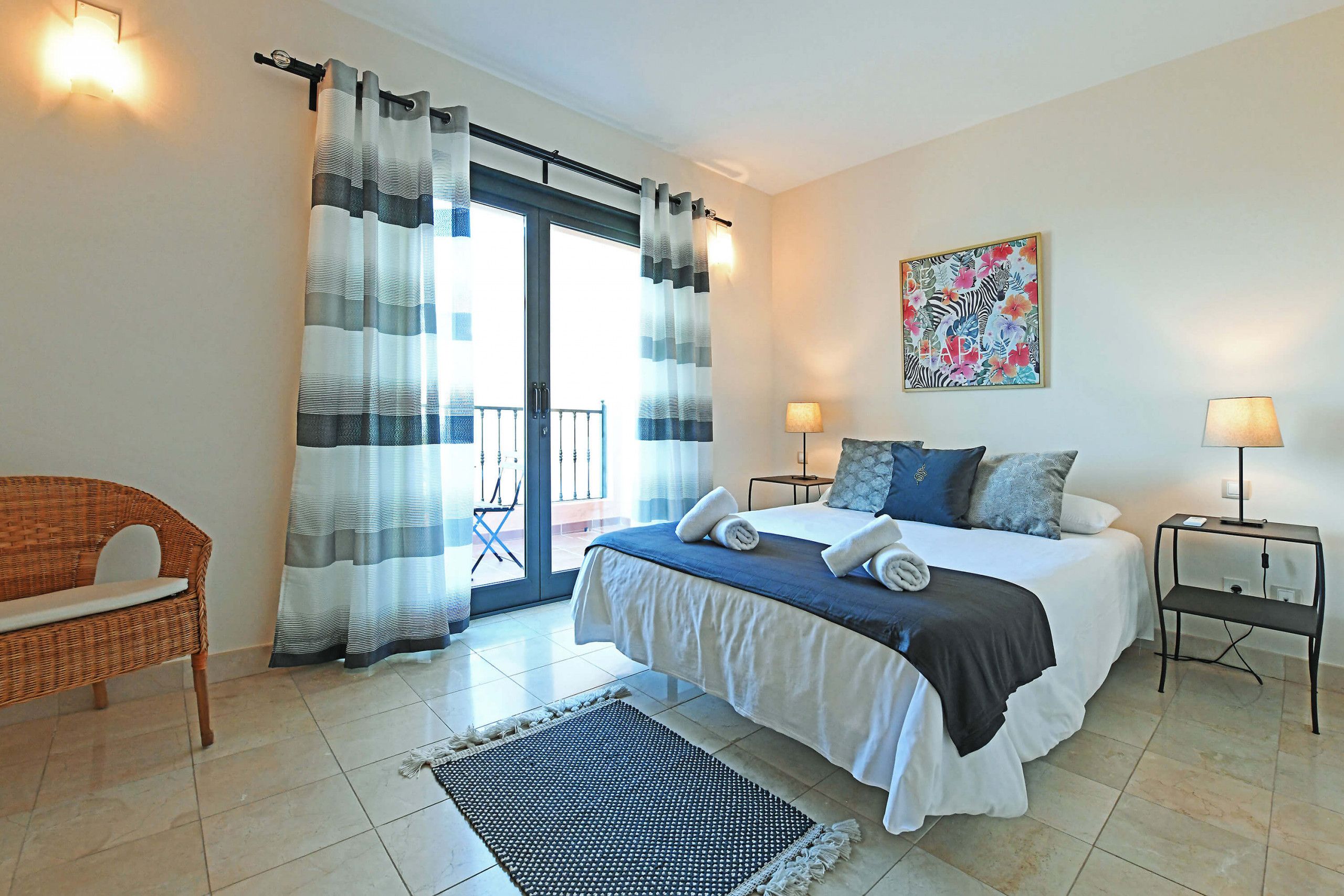 A bedroom with a bed, two bedside tables, a chair, a large window with curtains, and a wall-mounted artwork.