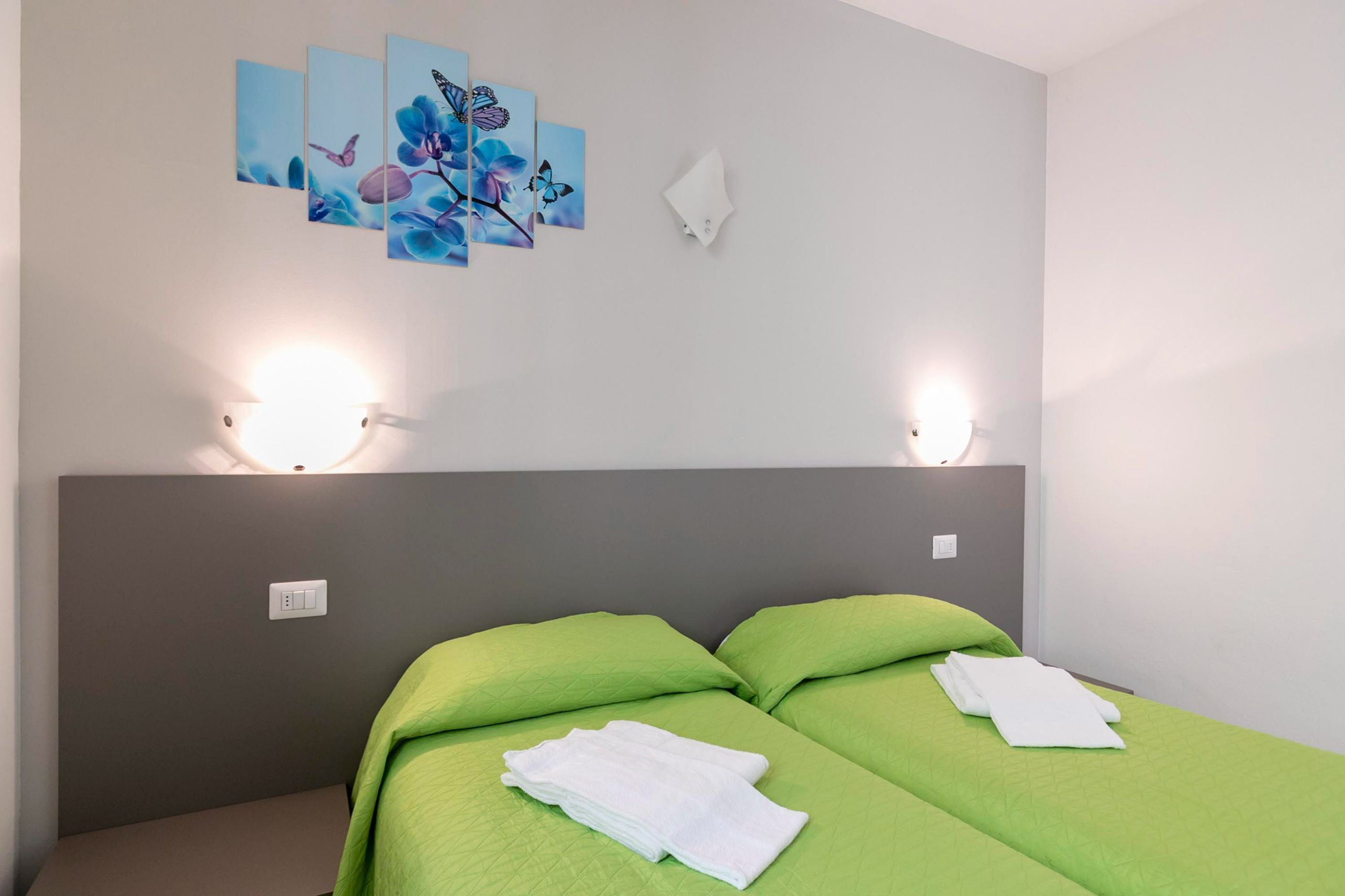 Une chambre d’hôtel avec deux lits, une literie verte, un luminaire fixé au mur et un tableau représentant un papillon.
