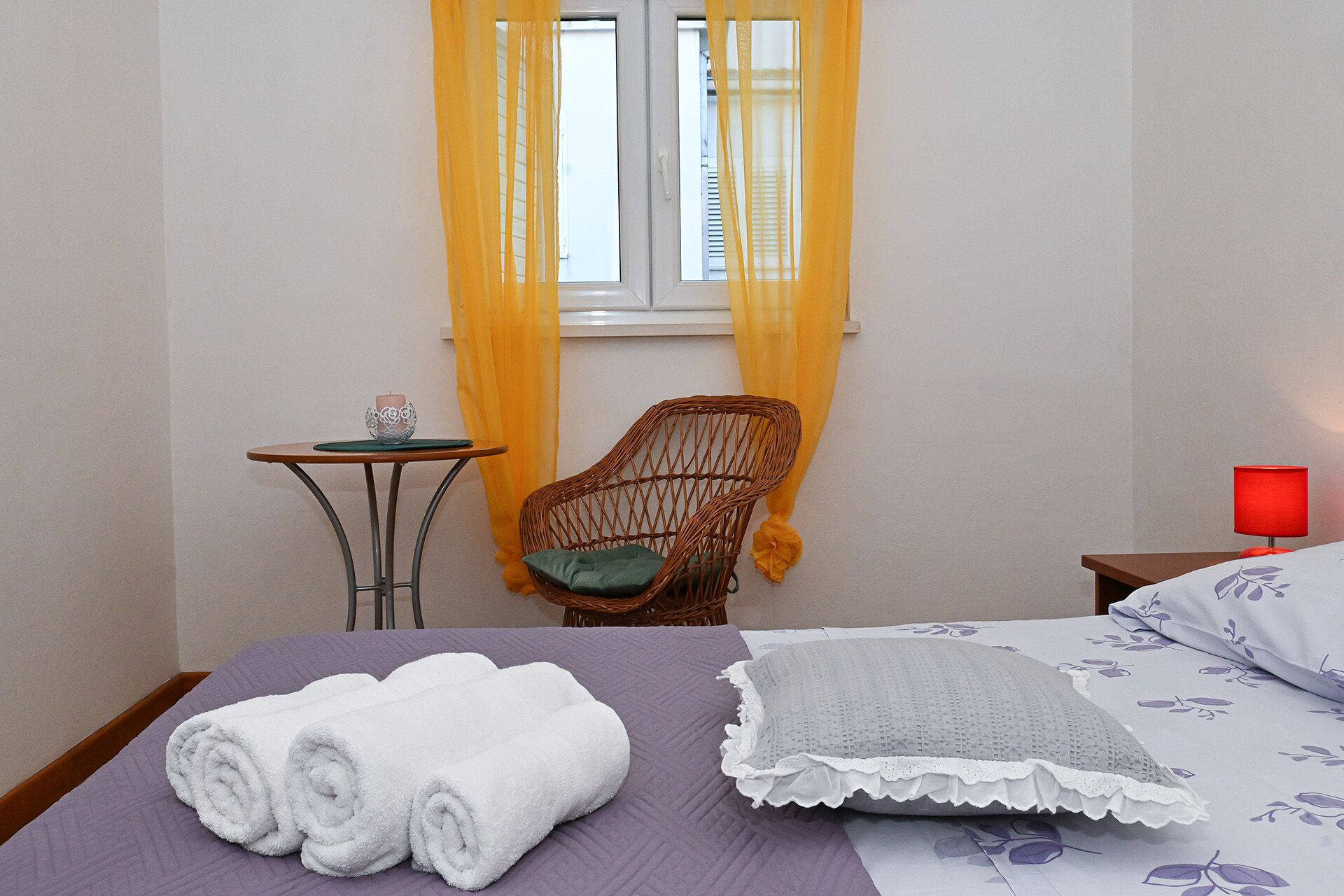 A hotel room with a bed, a chair, a small table, and a window with yellow curtains.