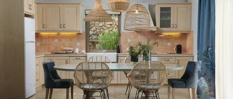 A modern kitchen with wooden cabinets, a central island with chairs, pendant lights, and a view of greenery outside.