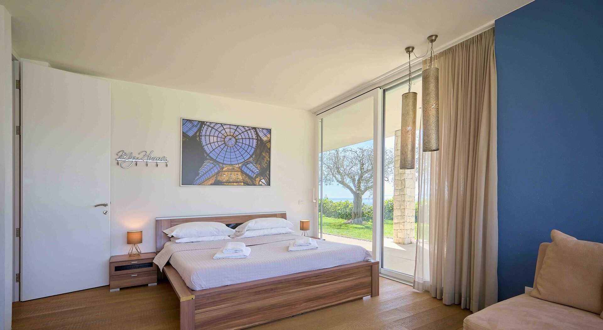 A bedroom with a bed, a nightstand, a wall-mounted artwork, a sliding glass door, and a view of greenery outside.