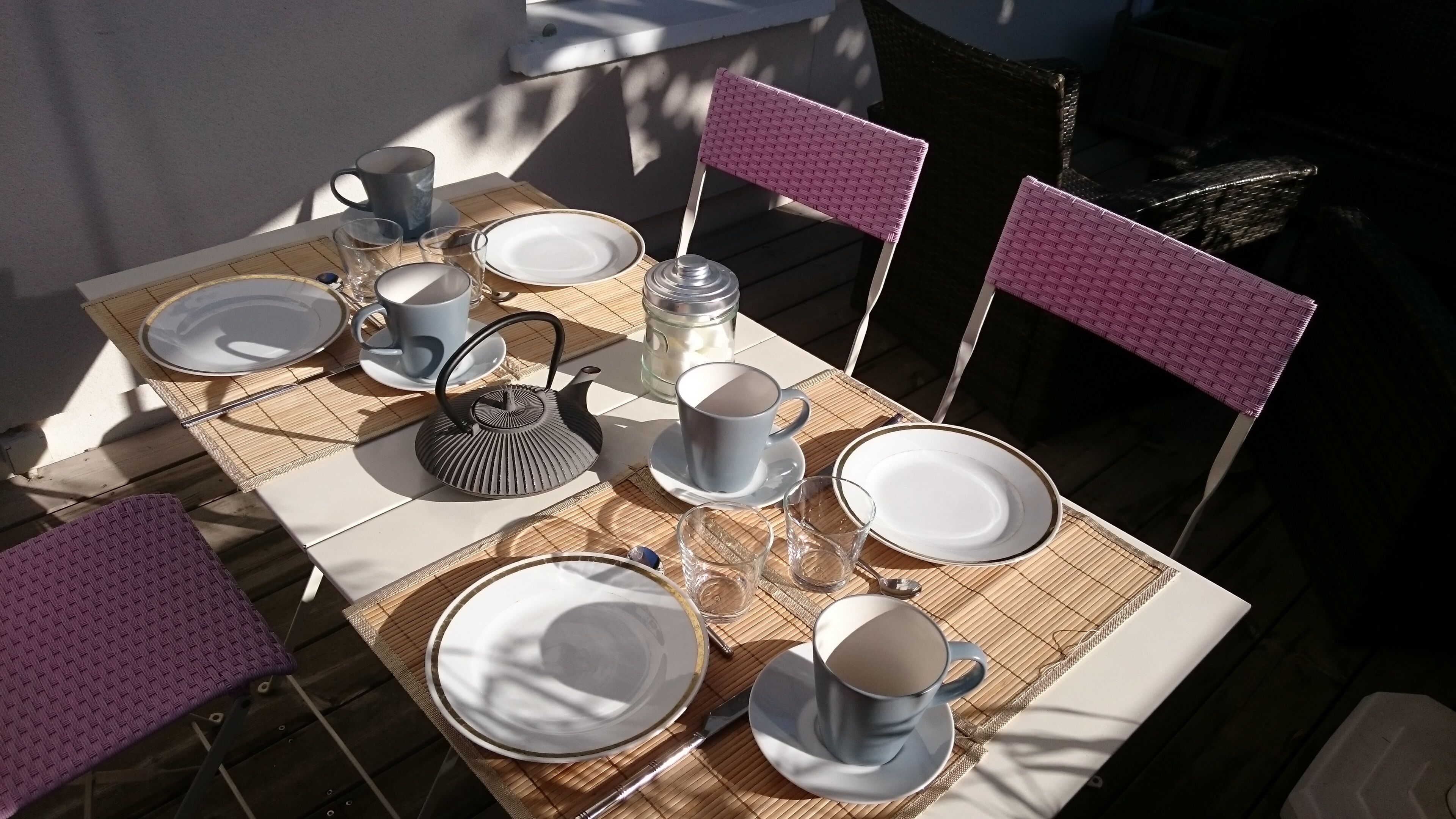 A table set with plates, cups, and a kettle, surrounded by chairs with a woven seat.