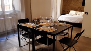A neatly set dining table with chairs, a bed with white linens, and a wooden headboard in a room with a plain wall and a window. - Prestigious "farnese loft" (Roma)
