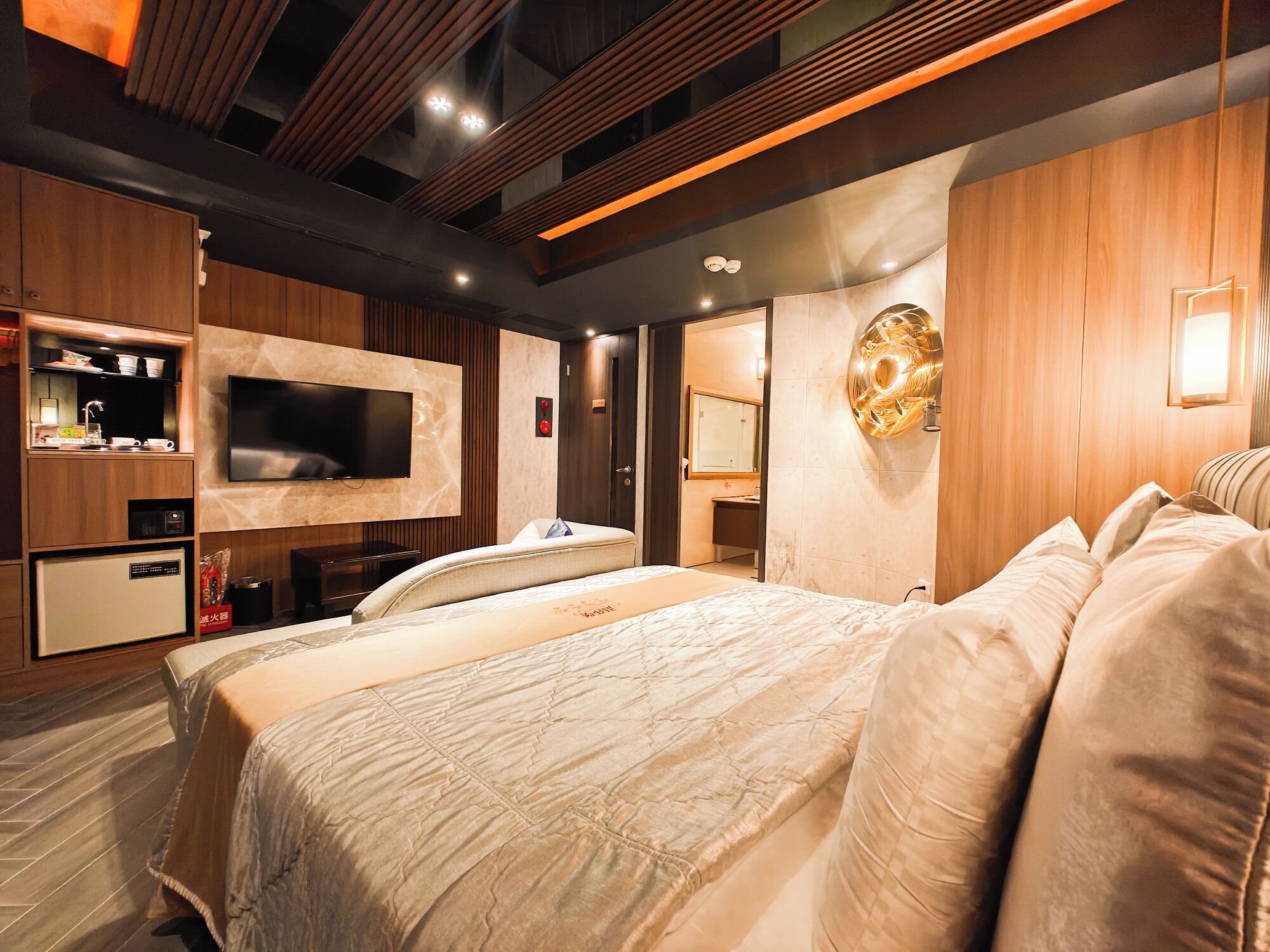 A modern hotel room with a large bed, a flat-screen TV mounted on the wall, a small kitchenette, and a bathroom visible through an open door.