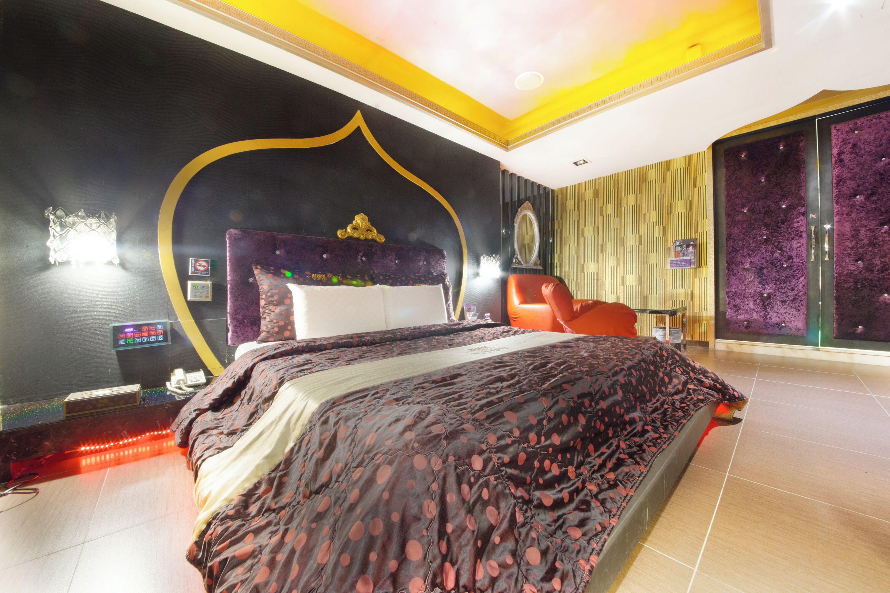 A modern bedroom with a large bed, a headboard with a decorative top, a red chair, and a wall-mounted TV.