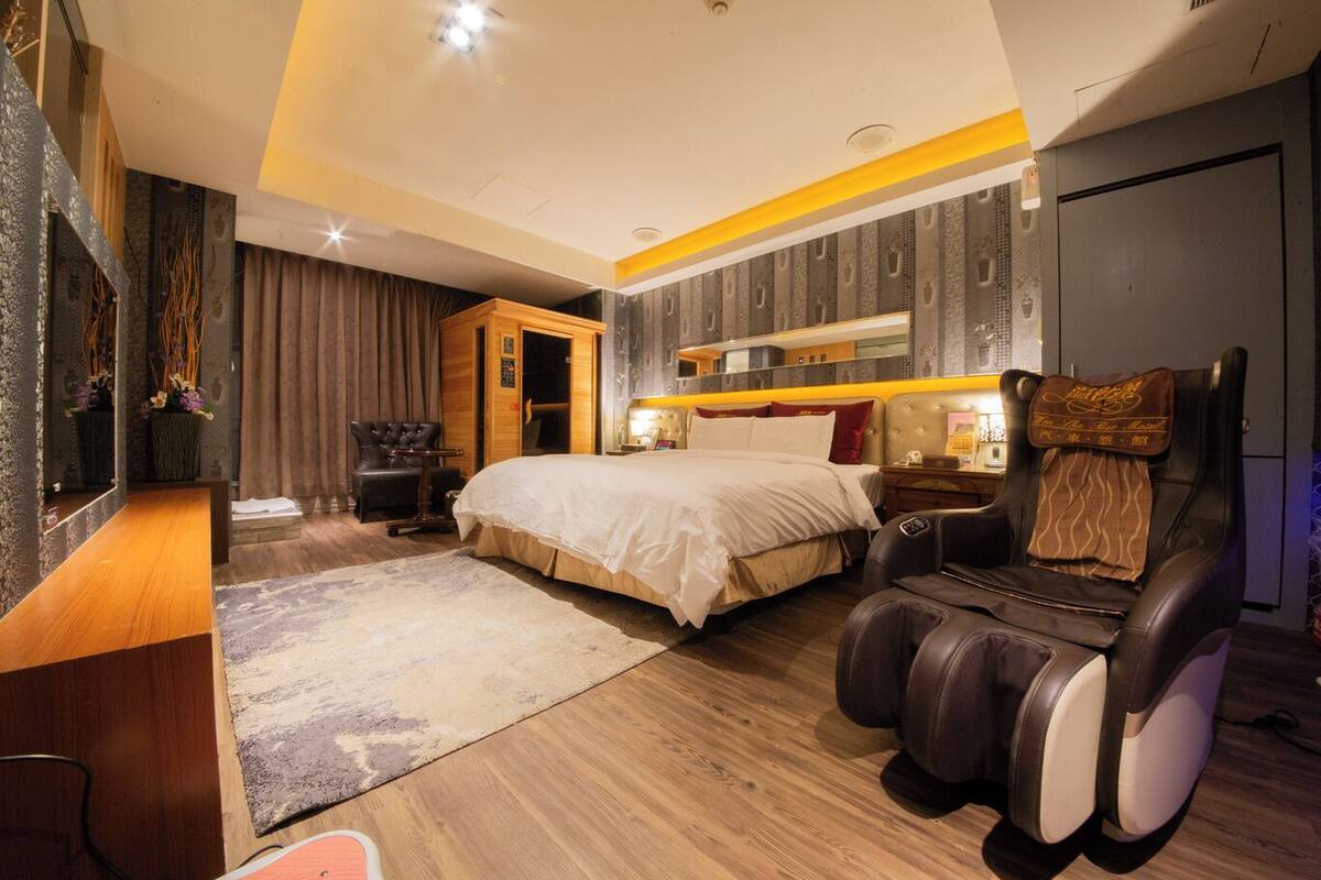 Una habitación de hotel moderna con una cama grande, una silla de masajes, un tocador con espejo y una zona de estar con una silla y una pequeña mesa.