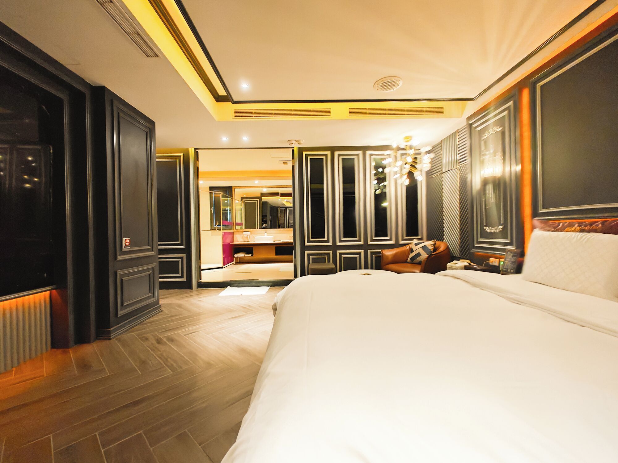 Una habitación de hotel con una cama grande, una zona de estar con sofá, un tocador con espejo, y un baño visible a través de una puerta abierta.