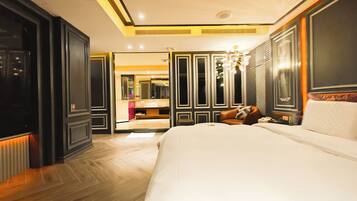 Una habitación de hotel con una cama grande, una zona de estar con sofá, un tocador con espejo, y un baño visible a través de una puerta abierta.