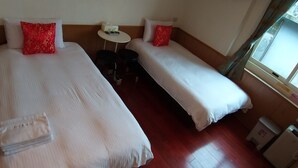 Een hotelkamer met twee eenpersoonsbedden, elk met een rood kussen en witte beddengoed. Er is een klein rond tafeltje met een stoel, een prullenbak en een verwarming op de vloer.