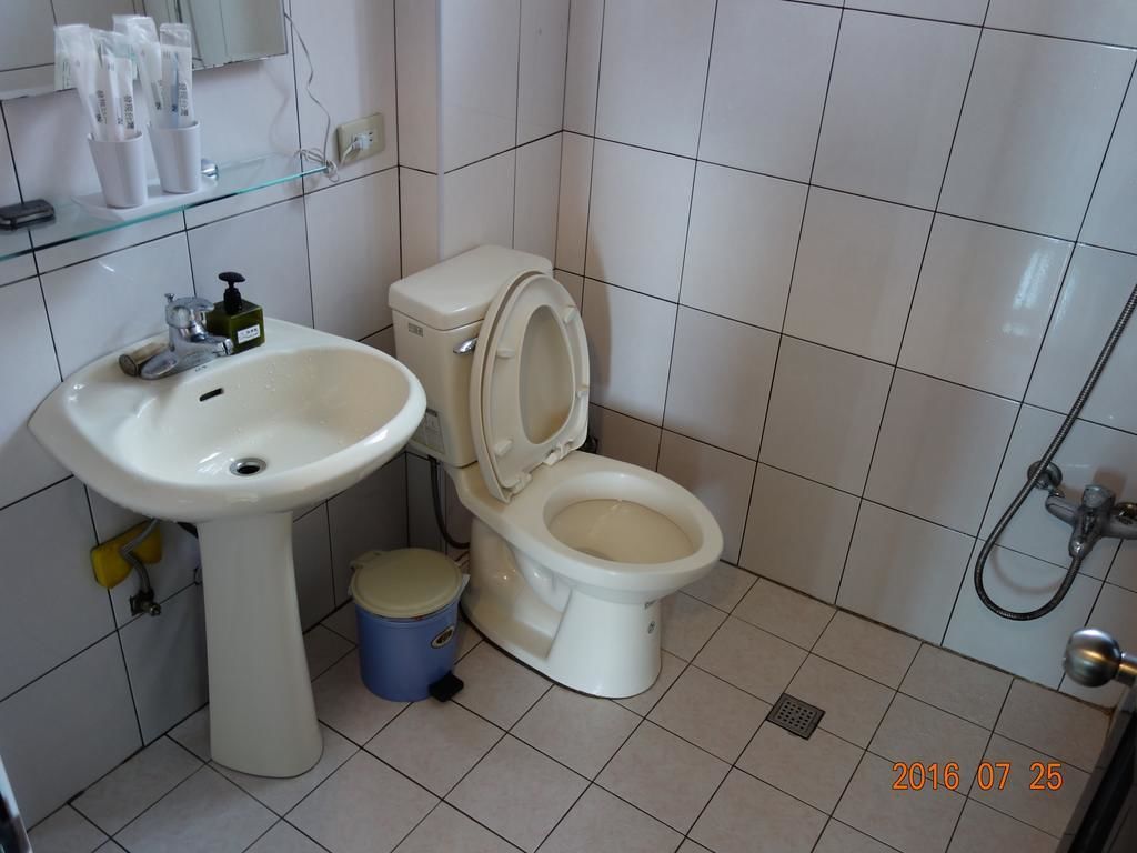 Ein Badezimmer mit einem weißen Waschbecken, einer Toilette und einer Dusche. Ein blauer Mülleimer und ein Glasregal mit Toilettenartikeln sind ebenfalls vorhanden.