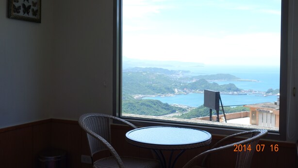 Een balkon met uitzicht op zee en heuvels, met rieten stoelen, een klein tafeltje en een vuilnisbak.