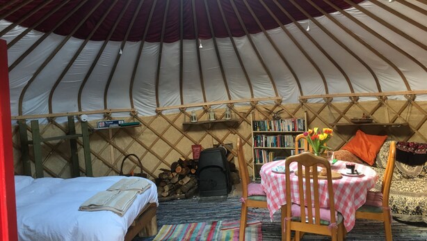 A yurt interior with a bed, a small table with chairs, a bookshelf, and a window. - Yurt [sleeps up to 6] & Bathhouse in E Cornwall UK - 5* Off-Grid  (Callington)