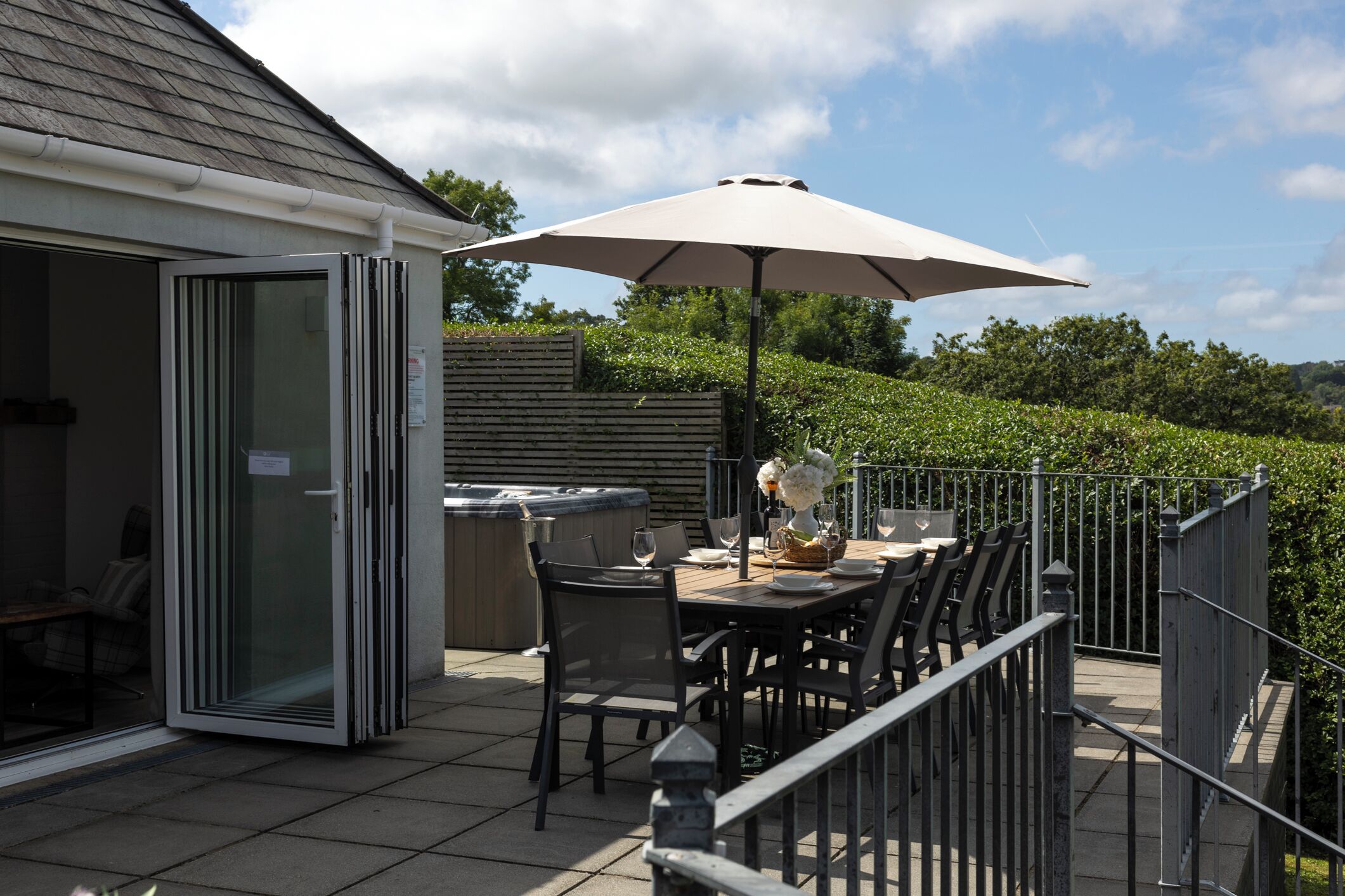Une terrasse extérieure avec une table dressée pour les repas, un jacuzzi et un grand parasol pour faire de l’ombre.
