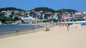 Una playa con gente caminando y tomando el sol, edificios al fondo y un cielo despejado.