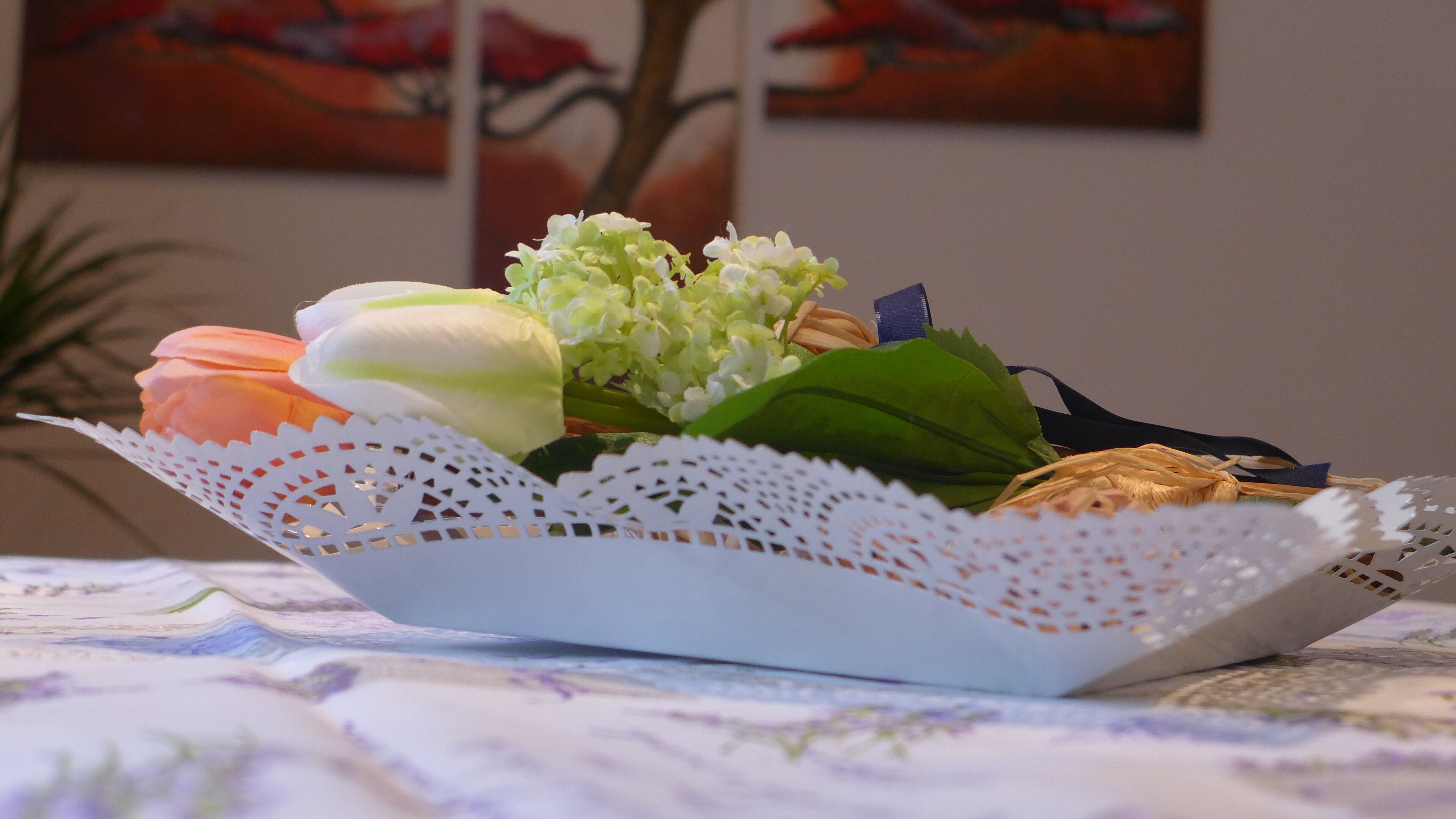 En blomsteropsætning i en hvid kurv på et bord med en blondeundertug.