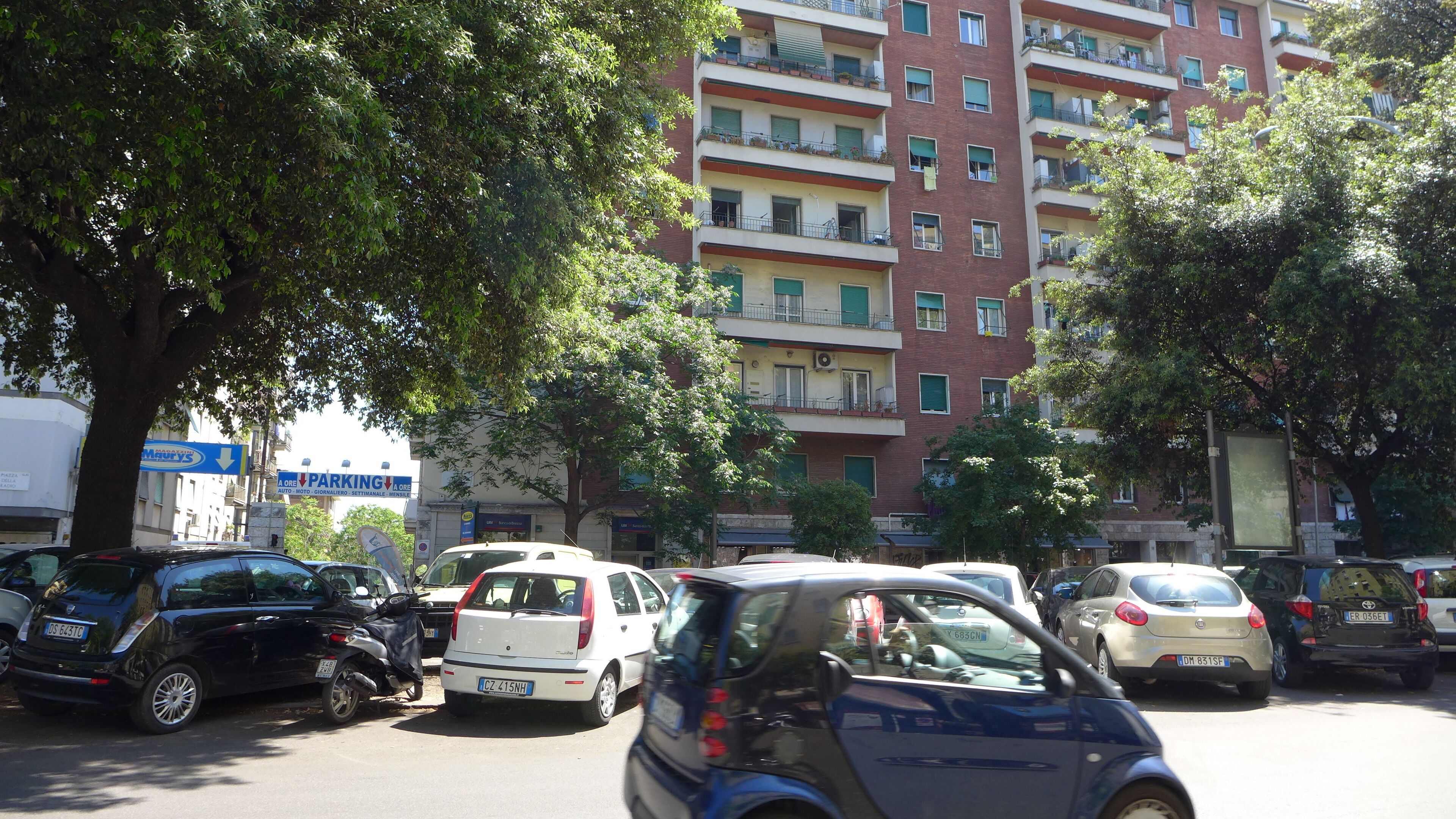 Un immeuble résidentiel à plusieurs étages, doté de balcons, avec des voitures garées et des arbres.