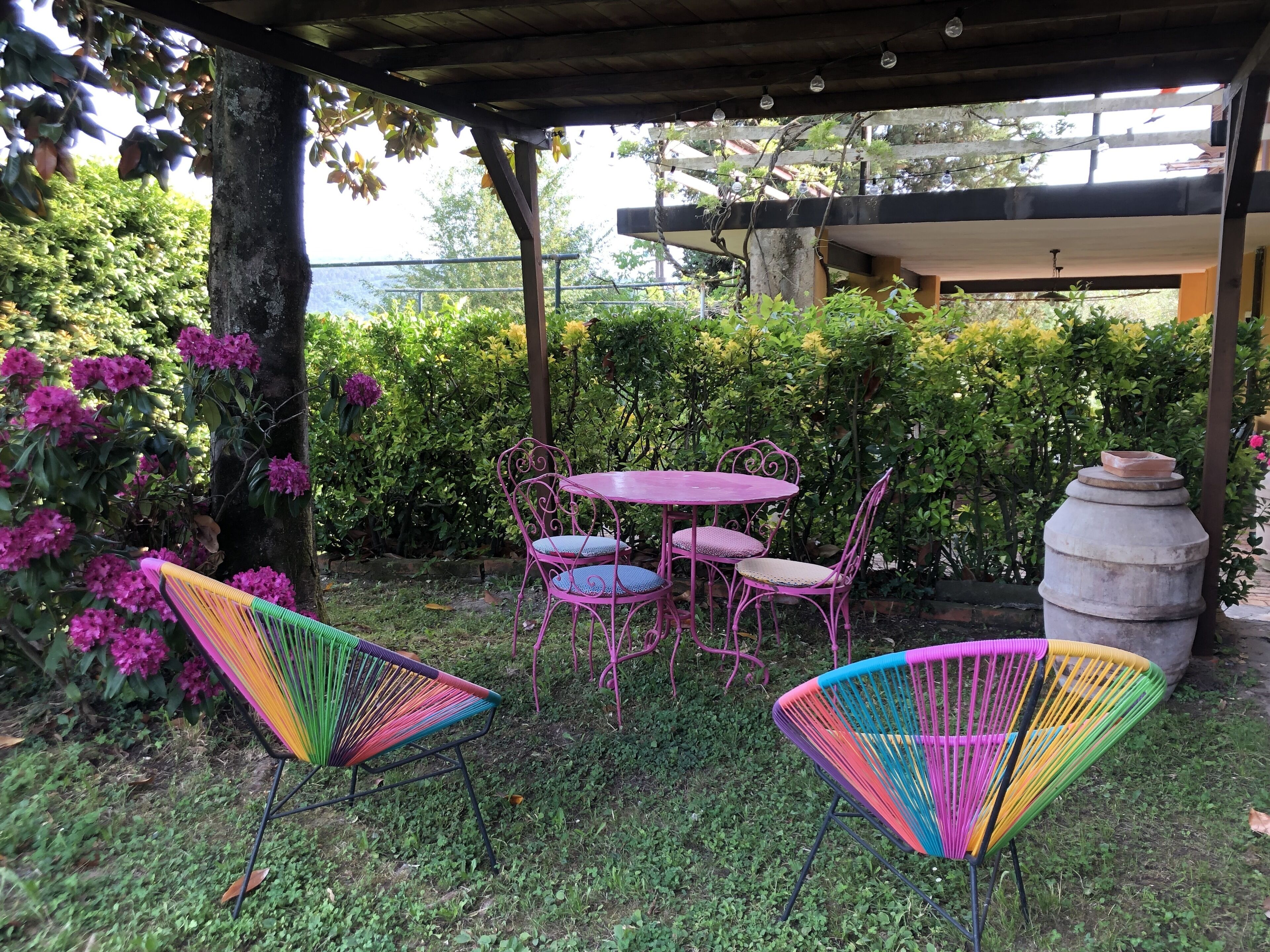 A covered outdoor seating area with a table and chairs, surrounded by lush greenery and flowering plants.