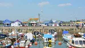 Ein Hafen mit vertäuten Booten, eine Pier mit Personen und verschiedene kleine Gebäude mit Schildern.