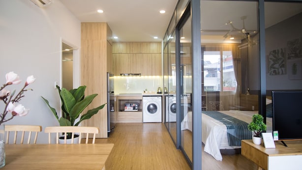 A modern hotel room with a wooden floor, a dining area with a table and chairs, a large potted plant, a bed with a view, a washing machine, and a television on a wooden side table. - Cuu Kim Son Hotel (Ho Chi Minh City)