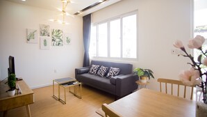A modern living room with a grey sofa, wooden coffee table, and a dining table. There are wall-mounted artworks and a potted plant on the windowsill. - Cuu Kim Son Hotel (Ho Chi Minh City)