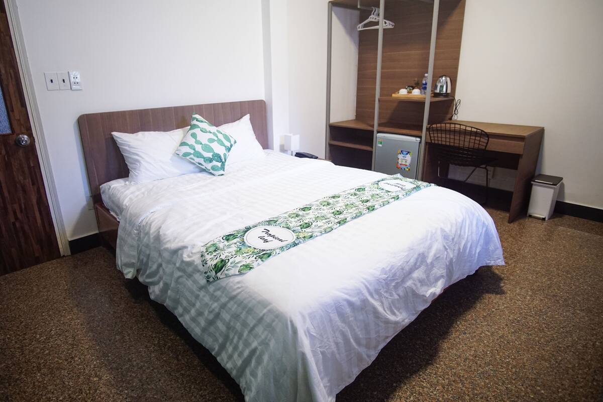 A neatly made bed with a floral bedspread, a wooden headboard, and a small desk with a chair and a trash bin.