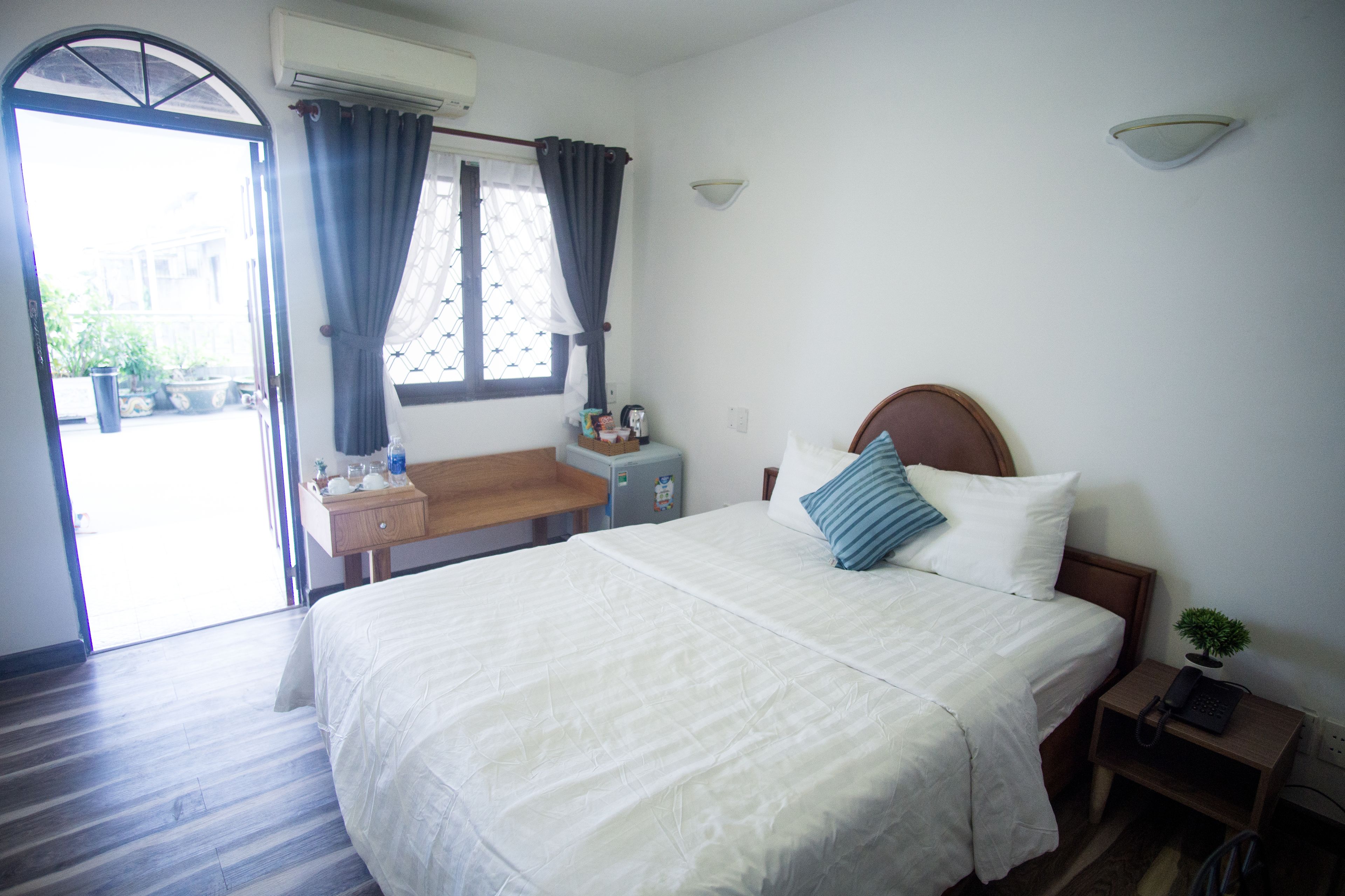 Habitación de hotel con cama, escritorio, silla, mesita, mesita de noche, planta en maceta y ventana con cortinas.