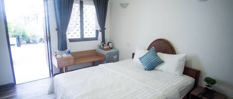 Habitación de hotel con cama, escritorio, silla, mesita, mesita de noche, planta en maceta y ventana con cortinas.