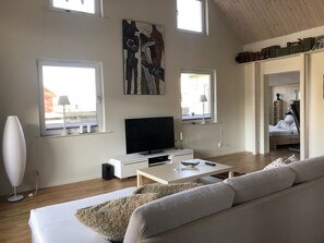 Un salon moderne avec un canapé, un téléviseur à écran plat et une table basse. Deux fenêtres laissent entrer la lumière naturelle, et une bibliothèque remplie de livres est visible au fond de la pièce.