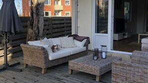 Een houten terras met een witte bank en een rieten voetenbankje, een boom en een gebouw op de achtergrond.