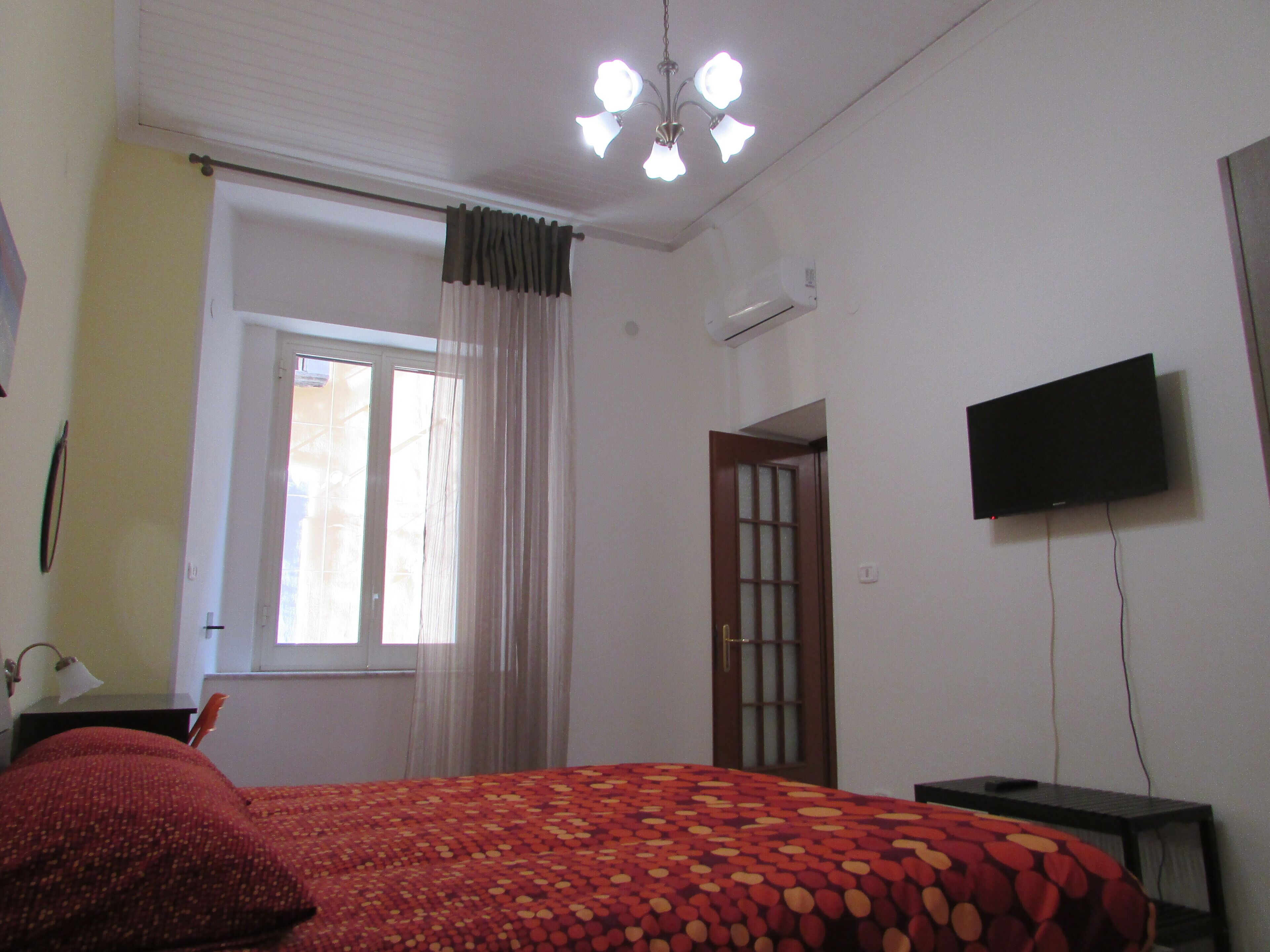 Un dormitorio con cama, televisor montado en la pared, ventana con cortinas y equipo de aire acondicionado.