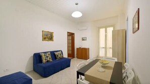 A living room with a blue sofa, a dining table with chairs, and a door leading to another room.
