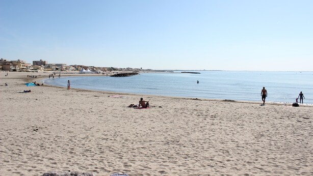 Une vaste plage de sable fin, une mer calme, quelques personnes et des bâtiments qui se dessinent à l’horizon.