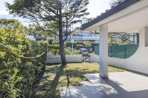 A covered outdoor area with a paved walkway, a tree, and a view of a residential building in the background.