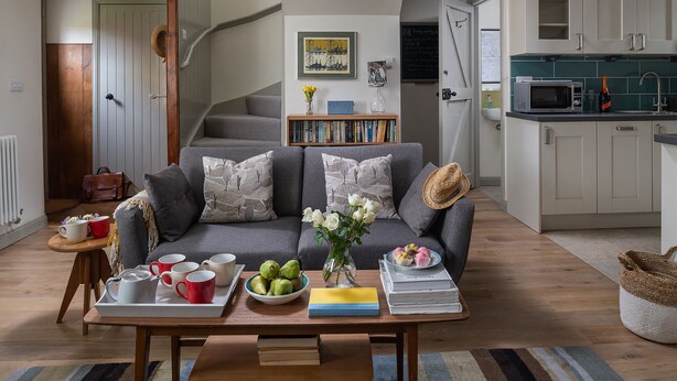 Un salon avec un canapé gris, une table basse avec des livres et un vase de fleurs, un escalier et un coin cuisine doté de meubles blancs et d’un four à micro-ondes.