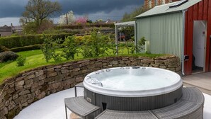 Un bain à remous avec un mur en pierre et des meubles en osier, entouré d’un jardin et, au fond, d’un bâtiment rouge.