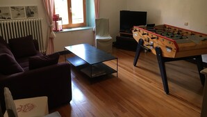 Ein Wohnzimmer mit einer Couch, einem Tisch, einem Kicker, einem Fernseher und einem Fenster mit Vorhängen.