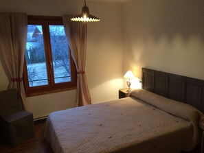 A bedroom with a bed, a nightstand with a lamp, a window with curtains, and a view of the outside. - Large apartment in mountain house in Risoul - 05600 (Risoul)