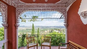 Uma varanda com colunas brancas ornamentadas, duas cadeiras de madeira e vista para uma paisagem verde com montanhas ao longe.