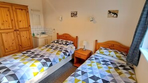 A bedroom with two beds, a wooden wardrobe, a small bedside table with a lamp, and a bathroom with a sink and mirror.