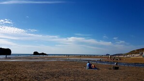 Uma praia com pessoas caminhando e sentadas, um afloramento rochoso na água e céu claro.