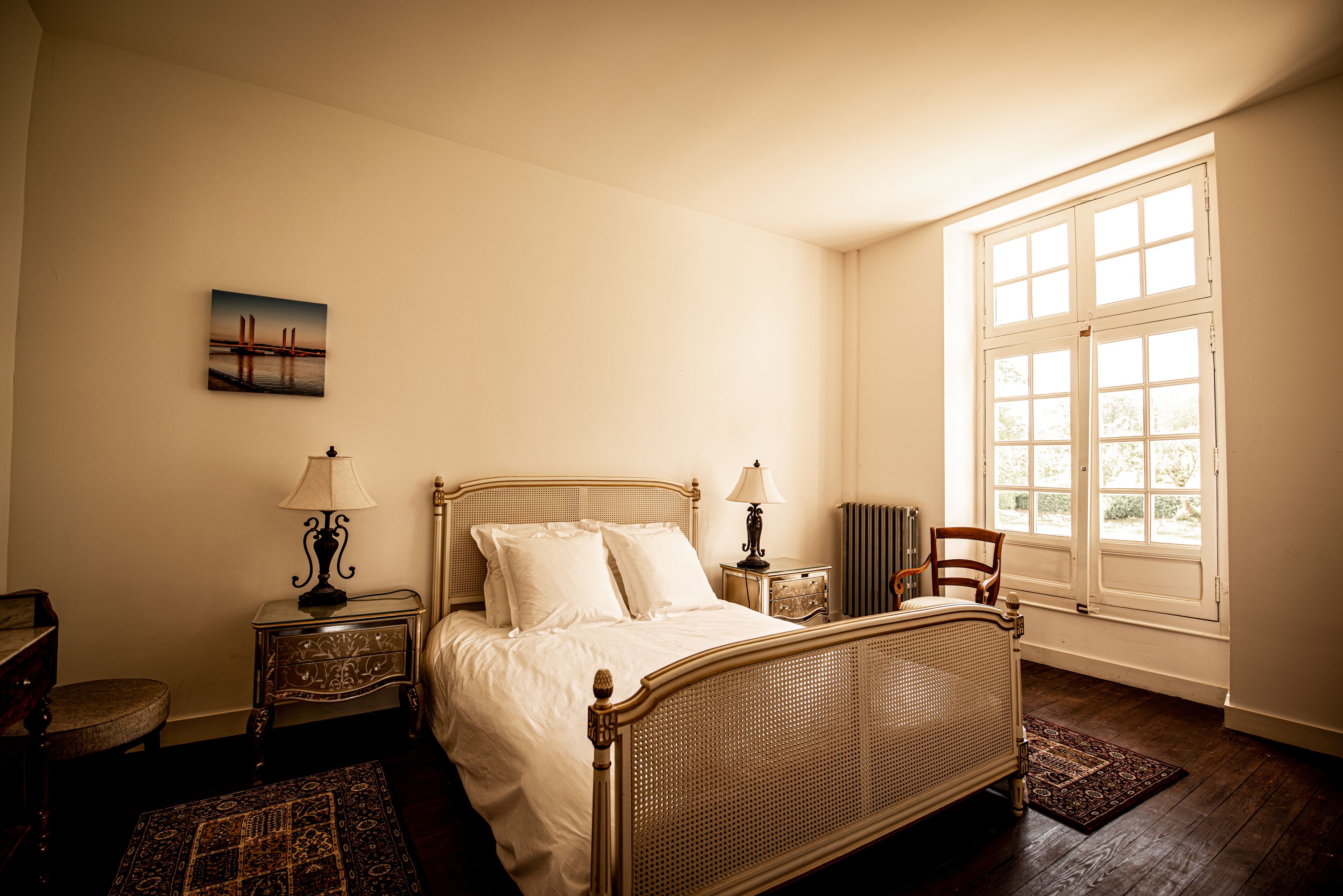 Een slaapkamer met een bed, twee nachtkastjes, een stoel, een raam en een schilderij aan de muur.