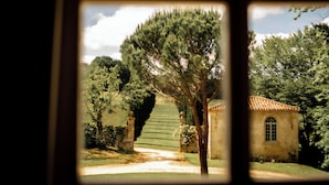 Une vue à travers la fenêtre d’un bâtiment donnant sur un jardin, un sentier et une petite construction surmontée d’un toit en tuiles.